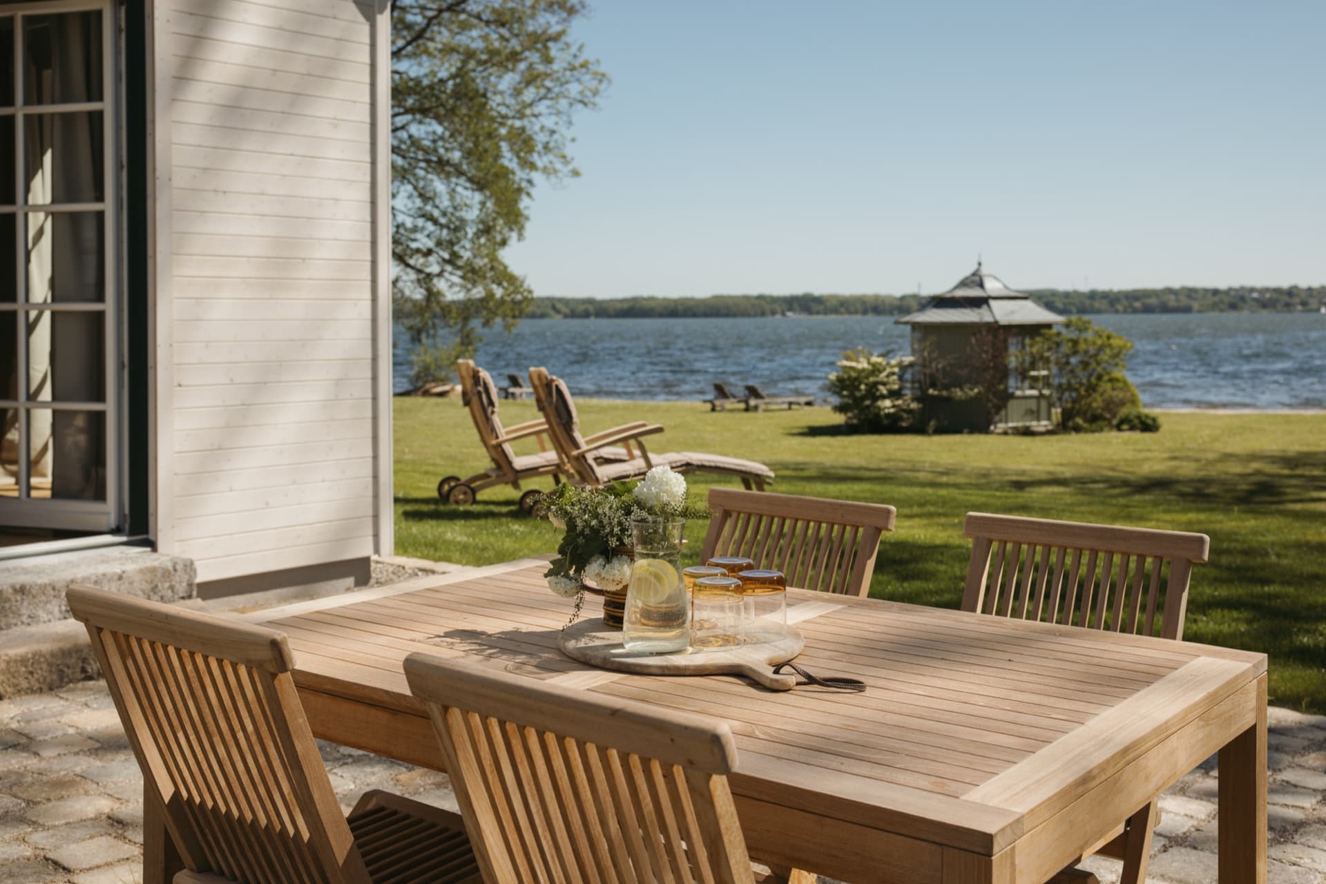 Terrasse mit Holztisch und Liegestühlen, Blick auf den See und den Pavillon